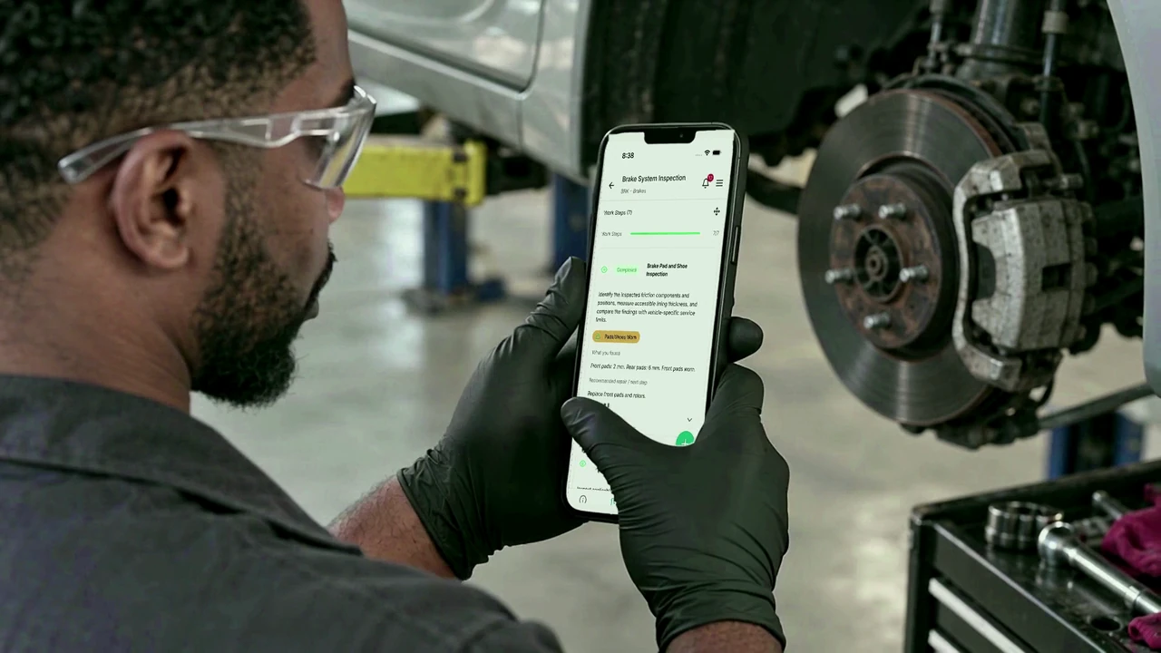 Technician Marcus reviews the brake inspection Work Steps on his iPhone beside the exposed brake rotor.